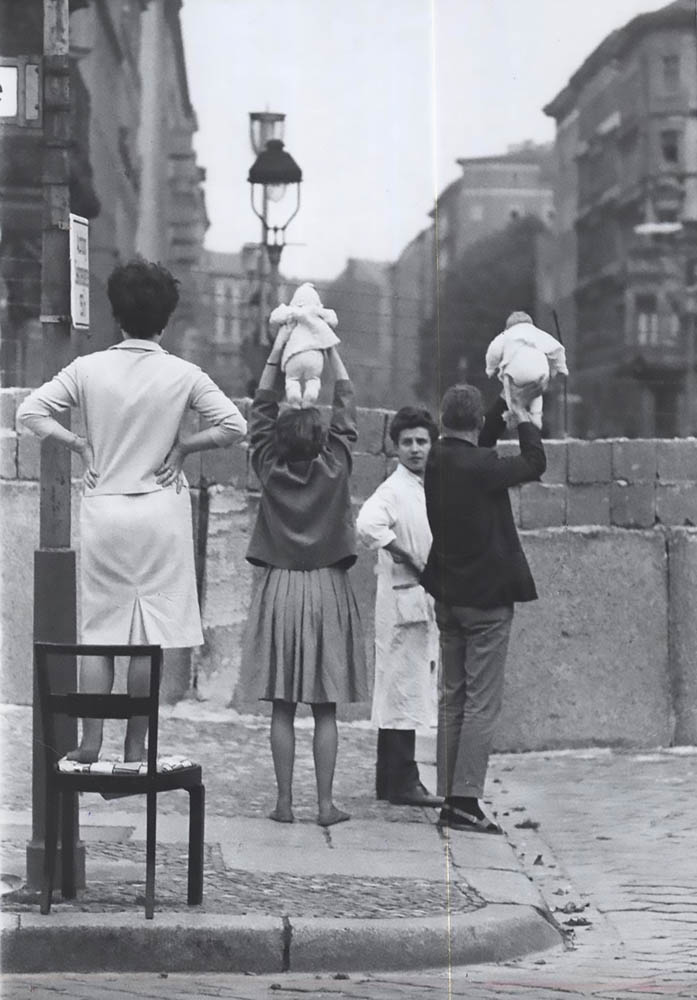 A small group of 4 people standing next to the Berlin wall. Two of these people are lifting babies above their head to show family members on the opposite side.
